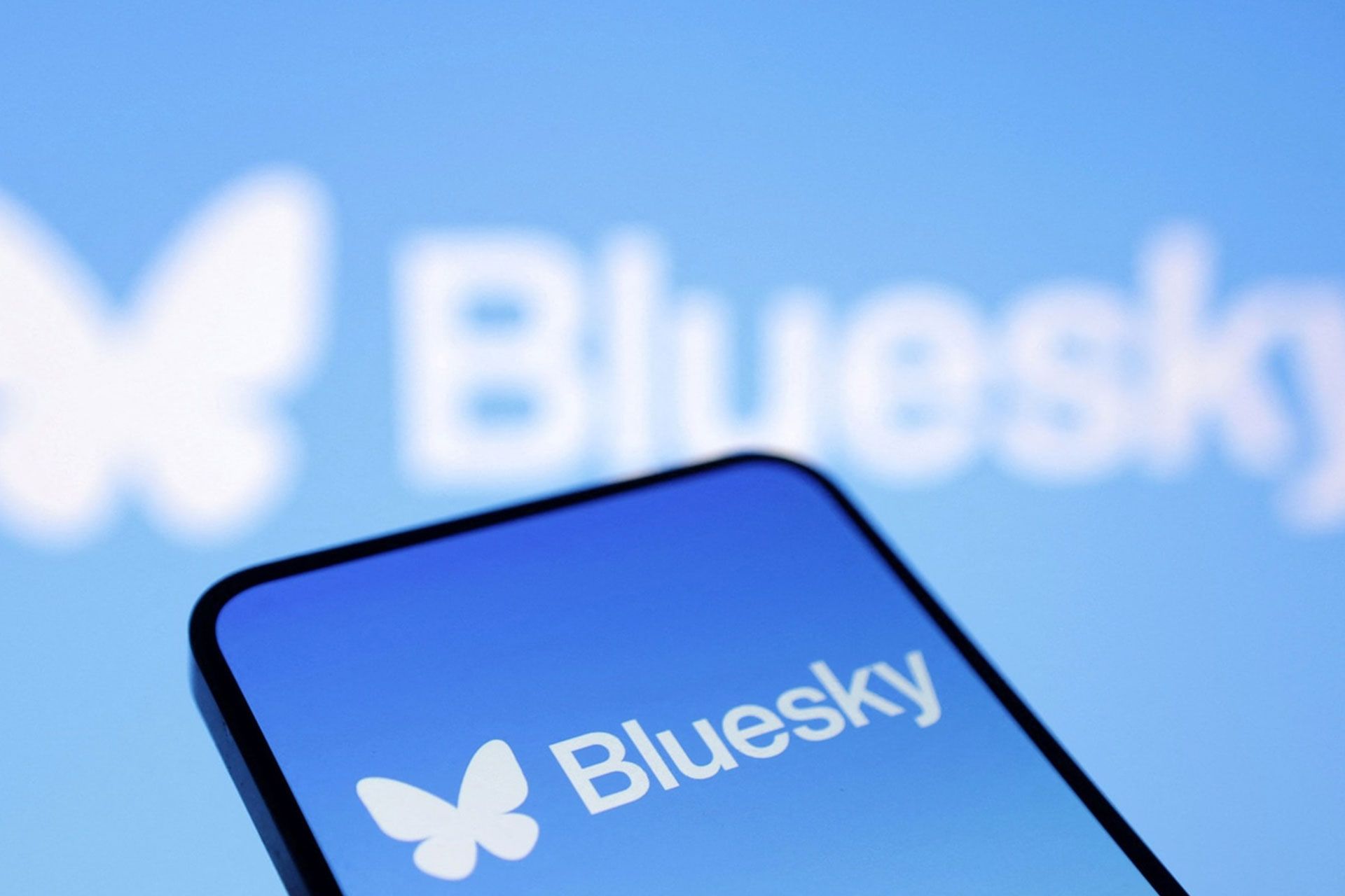bluesky-social-674a998120112d17060f7caa-1 لوگوی بلواسکای در نمایشگر گوشی روی پس زمینهی لوگو بلو اسکای روی دیوار